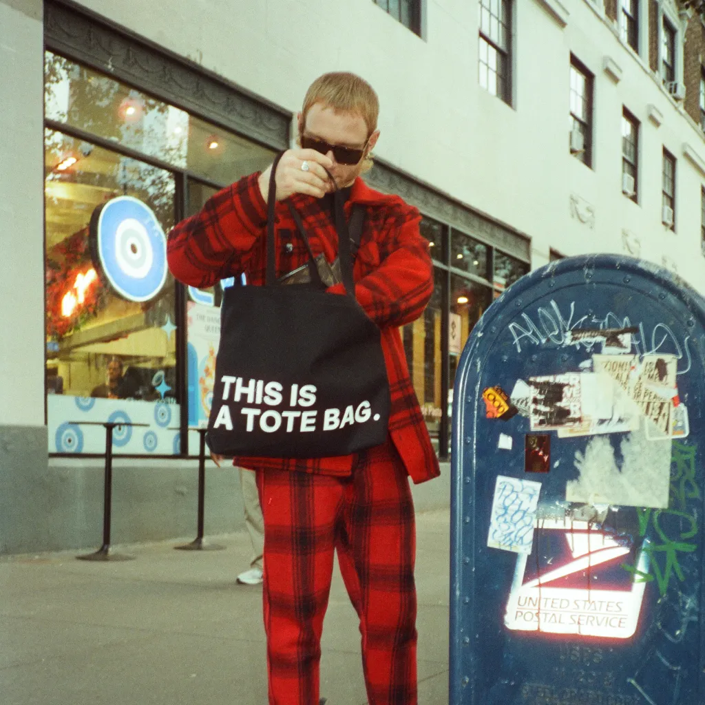 Man wearing red holding black tote bag that says THIS IS A TOTE BAG at mailbox