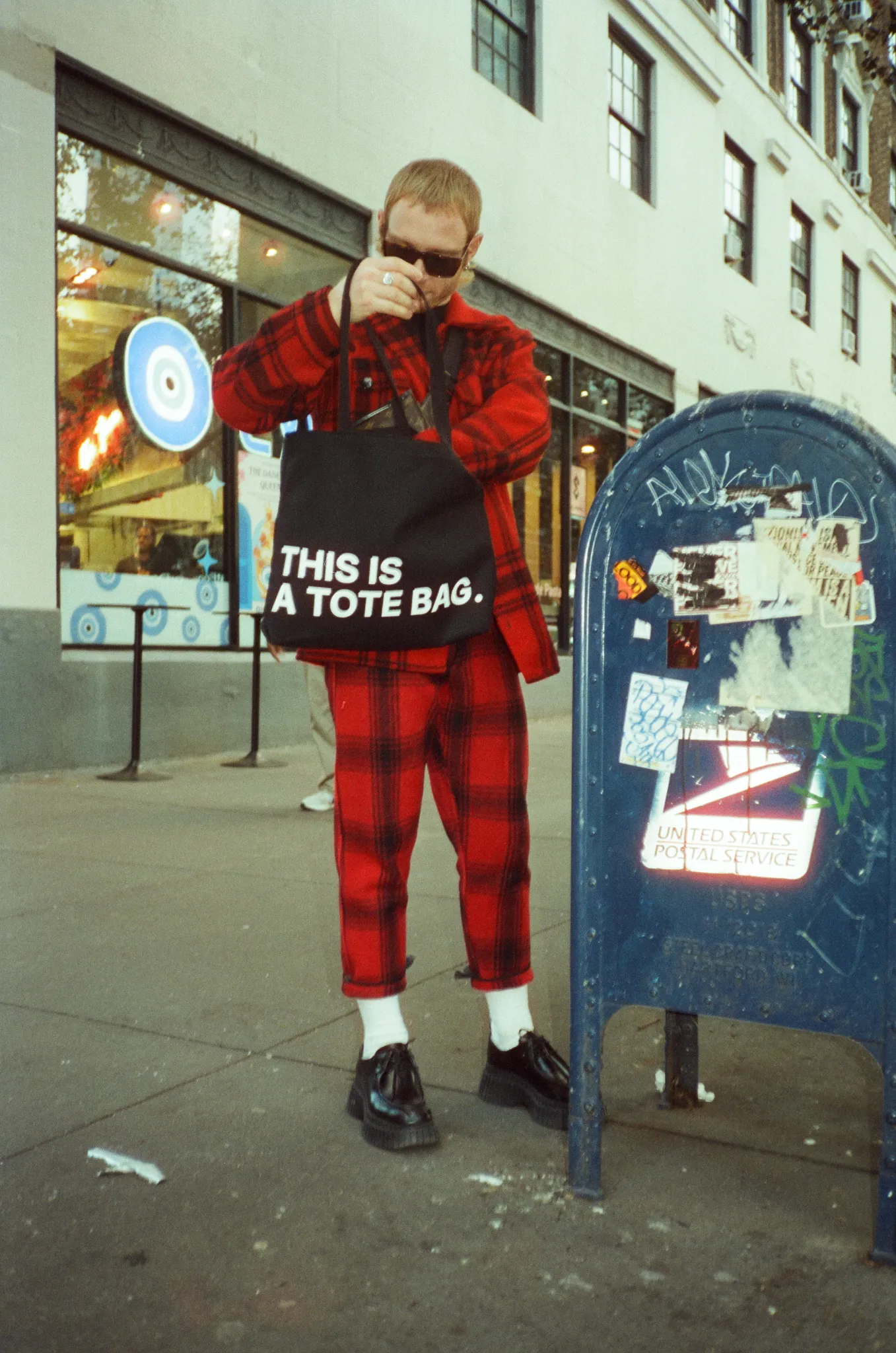 Man wearing red holding black tote bag that says THIS IS A TOTE BAG at mailbox